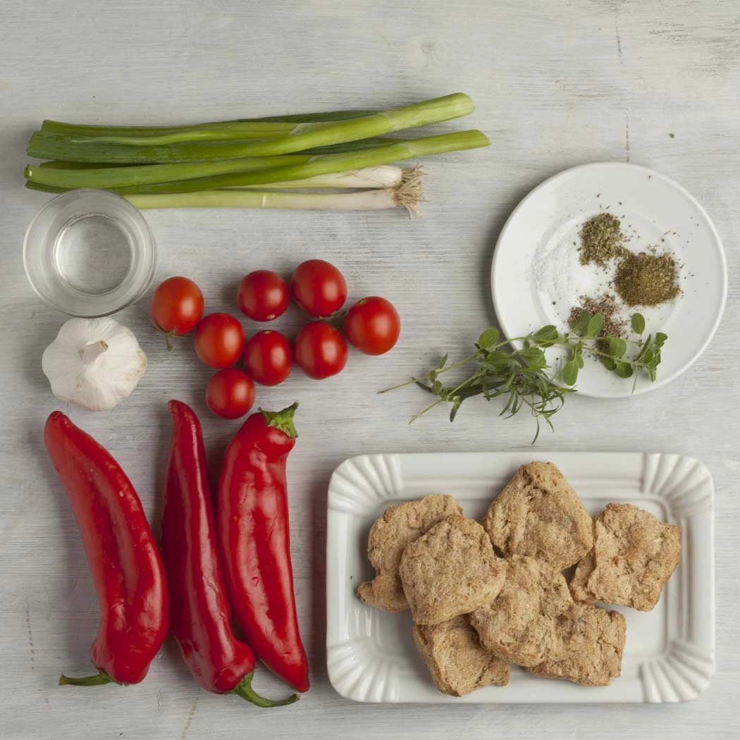 Zutaten zum Kochen: Sojaschnetzel, Spitzpaprika (Kapia), Tomaten, Zwiebel, Knoblauch, Öl und Gewürze auf einem hellen Tisch.