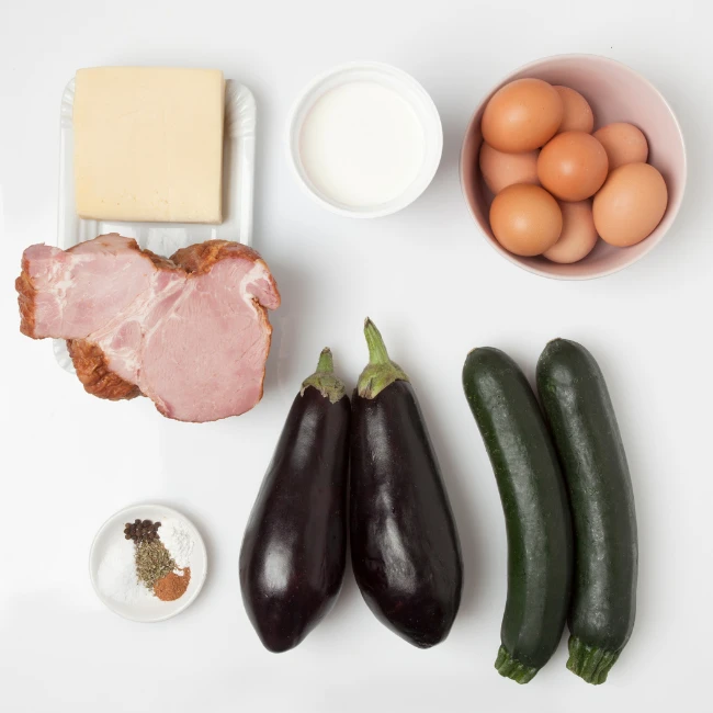 Ein Käseblock, Milch, Eier, geräuchertes Fleisch, Auberginen, Zucchini und eine Schüssel mit Gewürzen auf weißem Hintergrund.