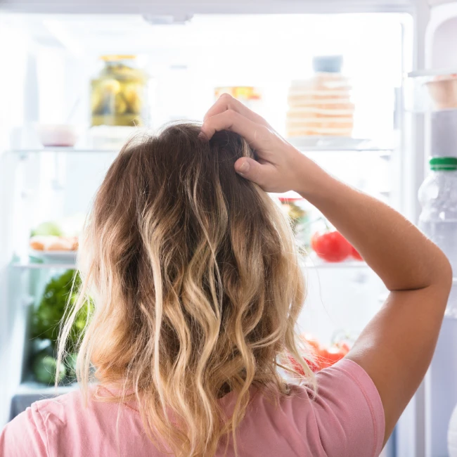 žena s vlnitými blond vlasmi stojí pred otvorenou chladničkou a s rukou na hlave premýšľa, aké zdravé jedlo si vybrať