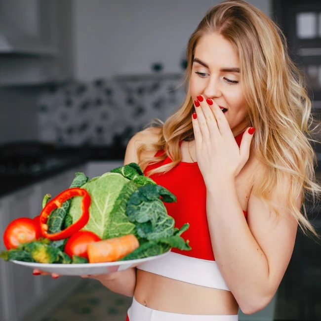 Štíhla mladá žena v červenom športovom tope prekvapene pozerá na tanier plný čerstvej zeleniny, ktorý drží v ruke