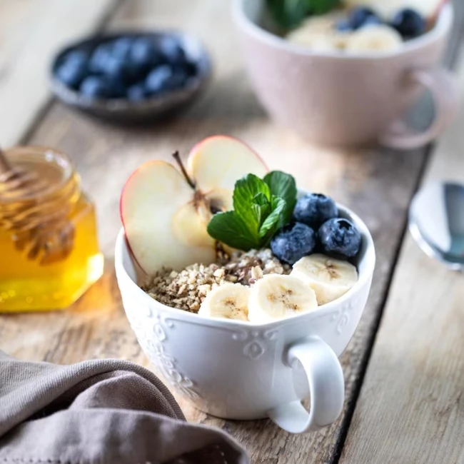 Zdravá raňajková kaša s kúskami banánu, jablka a čučoriedkami v bielej šálke s uškom