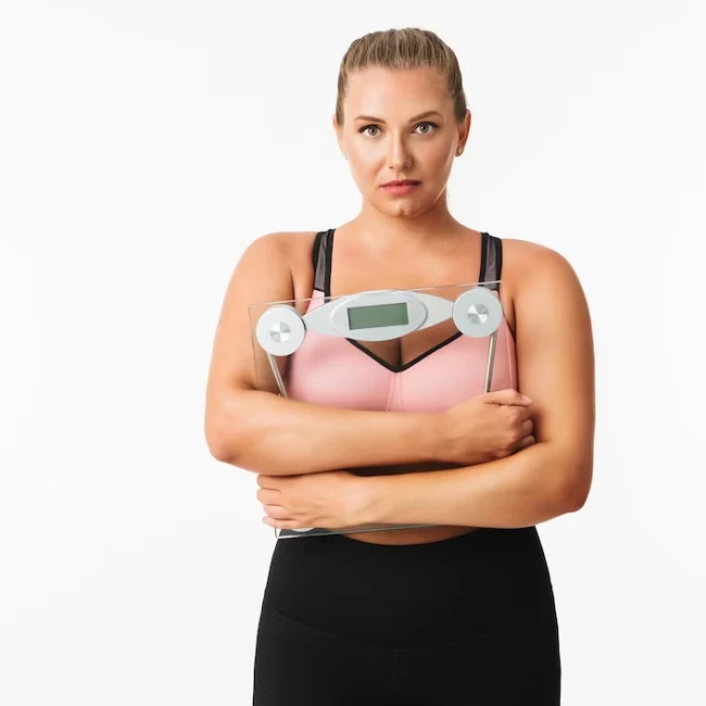 Nachdenkliche junge Frau in rosa BH und schwarzen Leggings hält eine ausgeschaltete Personenwaage in den Händen