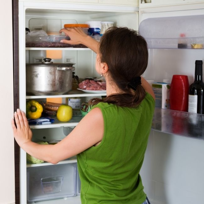 žena stojí pred otvorenou plnou chladničkou a vyberá jedlo z hornej poličky